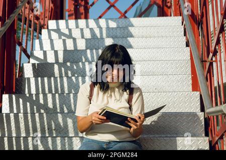 Vorbereitungs- und Universitätsstudium, junge lateinin, glücklich, ein Buch auf der Treppe zu lesen, sich darauf vorzubereiten, ein Profi zu werden. Stockfoto