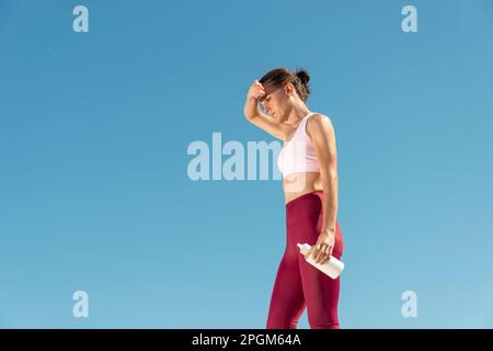 Fit sportliche Frau steht in der Sonne und hält eine Glasflasche Wasser, Hitzewelle Konzept. Stockfoto