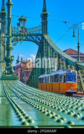 Die alte gelbe Straßenbahn, die auf der Freiheitsbrücke gegen die hellgrünen Metallkonstruktionen fährt, Budapest, Ungarn Stockfoto