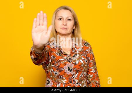 Weiße blonde Frau in einem gemusterten Kleid hebt ihre Hand in einem Stoppschild, isoliert auf gelbem Hintergrund. Stockfoto