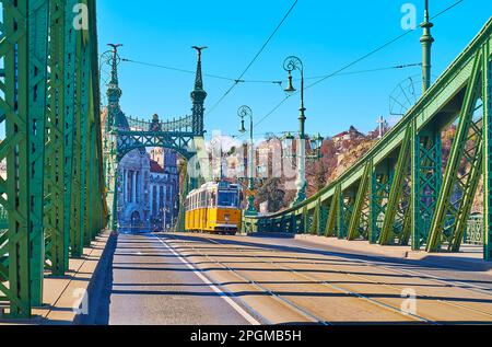 Die klassische gelbe Straßenbahn auf der Liberty Bridge gegen das Gebäude des Gellert Hotel and Baths in Buda, Budapest, Ungarn Stockfoto