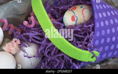 Nahaufnahme zeigt Ostereier, Küken, weiße Eier und Polkadot-Spielzeugeimer-Korb mit violettem, knisterndem Konfetti-Gras, das im Frühling auf einen Eierkarton gelegt wird. Stockfoto