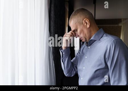 Porträt eines Mannes, der neben dem Fenster steht und seinen Kopf mit einer Hand hält, Kopfschmerzen hat oder aufmerksam ist. Stockfoto