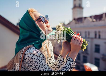 Stilvolle junge Frau mit grünem Retro-Schal und Sonnenbrille, die Frühlingsblumen riecht und die Stadtarchitektur genießt. Klassische Outdoor-Mode im Vintage-Stil Stockfoto