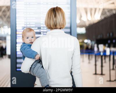 Mutter reist mit Kind, hält seinen kleinen Jungen am Flughafenterminal, überprüft Flugplan, wartet auf das Einsteigen in ein Flugzeug. Das Konzept „Reisen mit Kindern“ Stockfoto