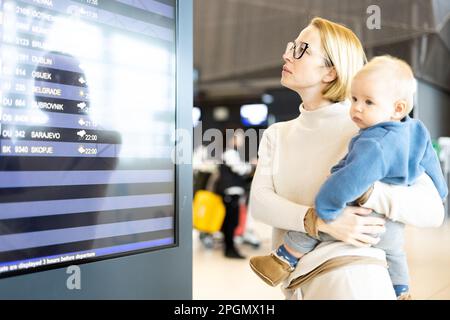 Mutter reist mit Kind, hält seinen kleinen Jungen am Flughafenterminal, überprüft Flugplan, wartet auf das Einsteigen in ein Flugzeug. Das Konzept „Reisen mit Kindern“ Stockfoto