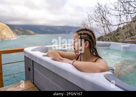 Nehmen Sie sich Zeit für sich. Badesachen im Freien mit Blick auf die Berge und das Meer. Eine Frau im schwarzen Badeanzug entspannt sich im Hotelpool und bewundert die Aussicht Stockfoto