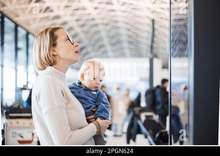 Mutter reist mit Kind, hält seinen kleinen Jungen am Flughafenterminal, überprüft Flugplan, wartet auf das Einsteigen in ein Flugzeug. Das Konzept „Reisen mit Kindern“ Stockfoto