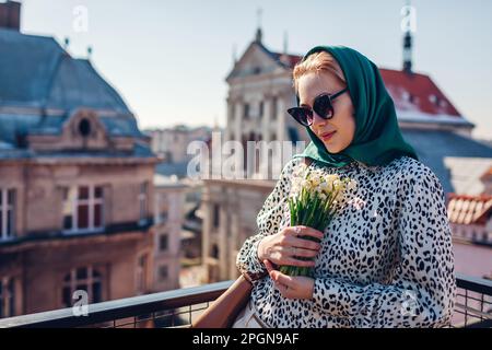 Stilvolle junge Frau mit grünem Retro-Schal und Sonnenbrille, die Frühlingsblumen hält und die Stadtarchitektur genießt. Klassische Outdoor-Mode im Vintage-Stil Stockfoto