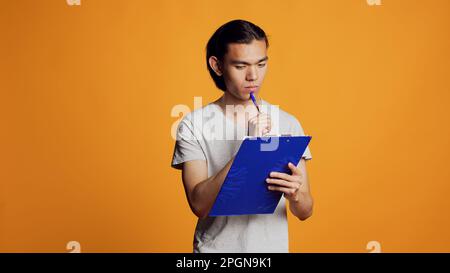 Selbstbewusster junger Mann, der Akten schreibt und sich Notizen macht, indem er Klemmbrett mit Papieren verwendet, um Informationen zu erhalten. Sorgenfreier, moderner Mensch, der Stift hält, um Dokumente im Studio zu unterschreiben, männliches Model. Stockfoto