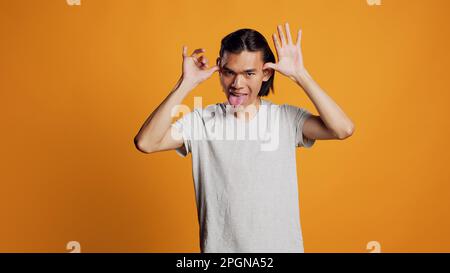 Amüsanter Kerl, der die Zunge rausstreckt und sich vor der Kamera komisch benimmt, junges sorgenfreies Model, das dumm und funky ist. Asiatischer Mann, der Comic-Momente genießt, herummacht und Spaß im Studio hat. Stockfoto
