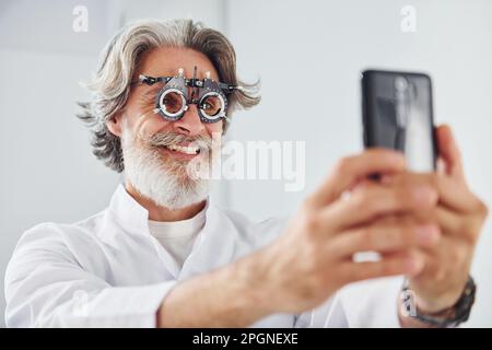 Ein älterer Mann mit grauem Haar und Bart ist in der Augenklinik und lächelt und hält ein Smartphone Stockfoto