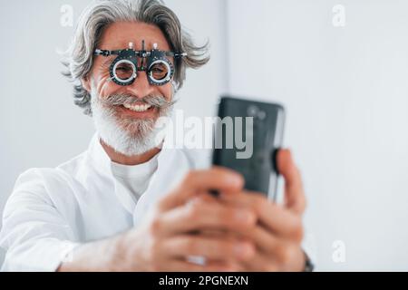 Ein älterer Mann mit grauem Haar und Bart ist in der Augenklinik und lächelt und hält ein Smartphone Stockfoto