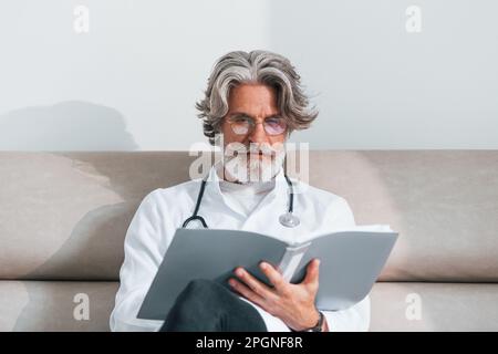 Leitender Arzt mit grauen Haaren und Bart in weißem Mantel ist in der Klinik sitzt auf dem Sofa mit Notizblock in den Händen Stockfoto