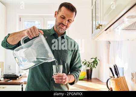 Mann, der Wasser aus einer Filterkanne in ein Glas in der Küche gießt Stockfoto