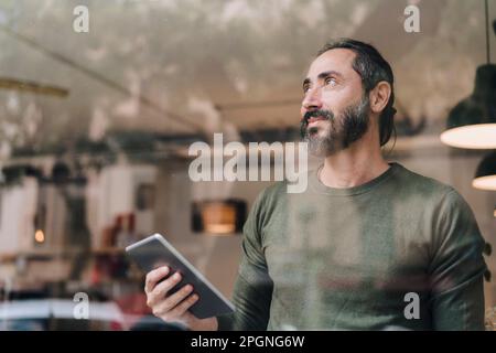 Durchdachter, reifer Mann mit Tablet-PC, der durch Glas gesehen wird Stockfoto