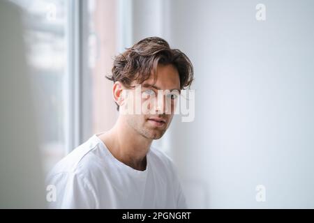 Lächelnder junger Mann vor weißen Wänden an Fenstern Stockfoto