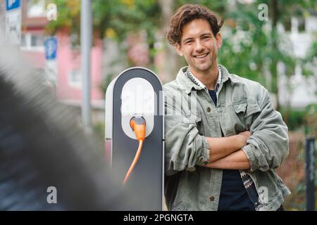 Glücklicher junger Mann, der sich auf ein Ladegerät für Elektrofahrzeuge stützt Stockfoto