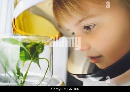 Ein Mädchen trägt einen Weltraumhelm und sieht sich eine kleine Pflanze an, die in einem Glasgefäß wächst Stockfoto