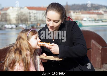 Die junge Mutter und ihre kleine Tochter saßen auf einer Bank am Ufer der Moldau, die Mutter fütterte ihre Tochter mit einem Sandwich, das sie bei einem lokalen Stockfoto