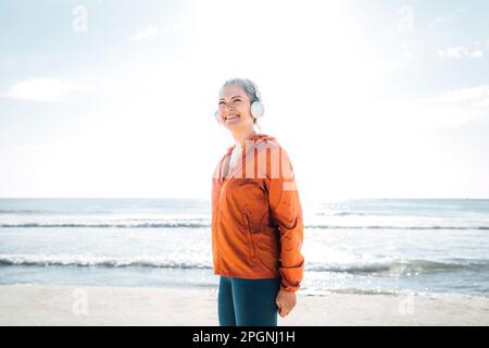 Lächelnde Erwachsene Frau mit Kopfhörern, die Musik am Strand hört Stockfoto