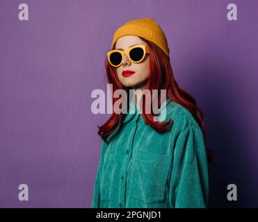 Rothaarige Frau mit Strickmütze und Sonnenbrille vor violettem Hintergrund Stockfoto