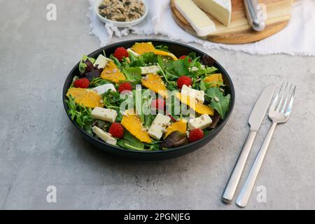 Frischer grüner Salat mit Brie-Käse auf schwarzem Teller auf grauem Tisch Stockfoto