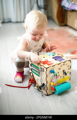 Das süße kleine Mädchen spielt draußen mit dem Busiboard auf grünem Gras. Pädagogisches Spielzeug für Kleinkinder. Das Mädchen öffnete die Tür, um den Brett zu würfeln. Stockfoto