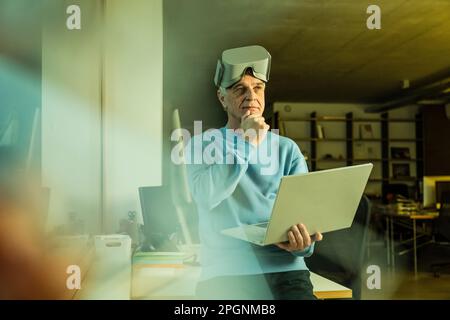 Aufmerksamer Geschäftsmann mit Hand am Kinn hält Laptop im Büro Stockfoto