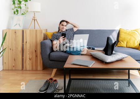 Junger Mann mit Beinprothese mit Smartphone, der zu Hause auf dem Sofa sitzt Stockfoto