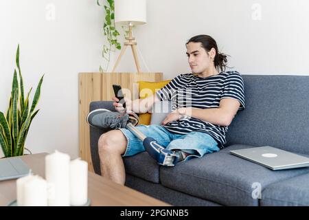 Junger Mann mit Beinprothese, der zu Hause ein Smartphone auf dem Sofa benutzt Stockfoto