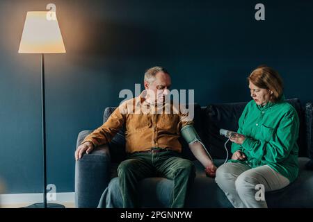 Seniorin prüft Blutdruck eines Mannes, der zu Hause auf dem Sofa sitzt Stockfoto