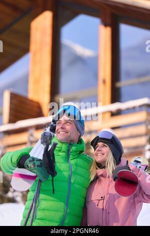 Lächelnder Mann und lächelnde Frau stehen mit Skiern vor dem Chalet Stockfoto