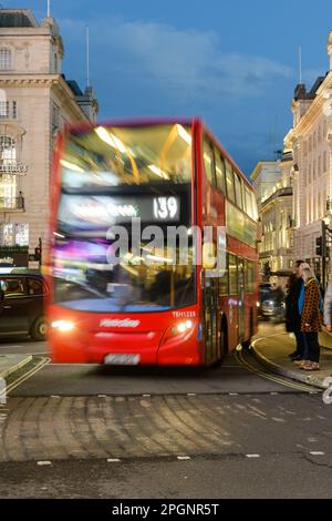 London, Großbritannien - 17. März 2023; verschwommener roter Londoner Bus fährt durch Piccadilly Circus mit Geschwindigkeit, während Fußgänger in der Abenddämmerung auf die Straße warten Stockfoto