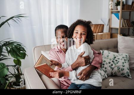 Glückliches Mädchen mit Buch, das mit Großmutter zu Hause auf dem Sofa sitzt Stockfoto