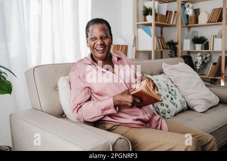 Glückliche Seniorin mit Buch, die zu Hause auf dem Sofa sitzt Stockfoto