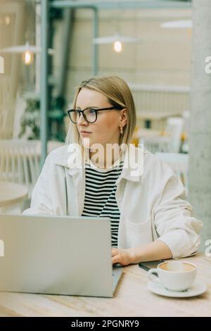 Nachdenkliche Geschäftsfrau mit Brille, die am Tisch im Café ein Notebook benutzt Stockfoto