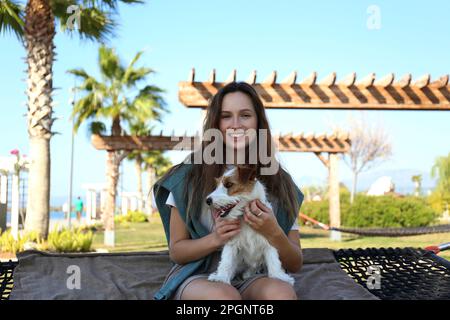 Glückliche junge Frau mit Hund, die auf einer Hängematte im Park sitzt Stockfoto