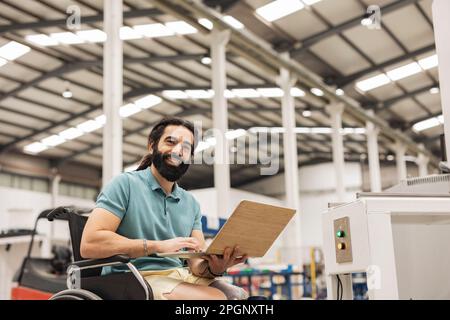 Glücklicher Ingenieur, der im Rollstuhl sitzt und in der Roboterfabrik an einem Laptop arbeitet Stockfoto
