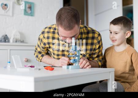 Ein lächelnder Junge, der neben dem Vater sitzt und durch das Mikroskop am Tisch schaut Stockfoto