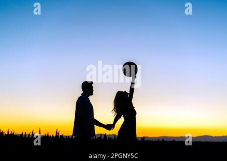 Silhouette-Mann, der bei Sonnenuntergang die Hand einer Freundin hält und auf dem Feld einen Hut hebt Stockfoto