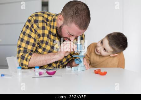 Süßer Junge, der Vater beobachtet, der durch das Mikroskop am Tisch schaut Stockfoto