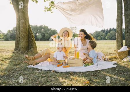 Glückliche Frauen sitzen mit Söhnen auf einer Picknickdecke und genießen den Park Stockfoto