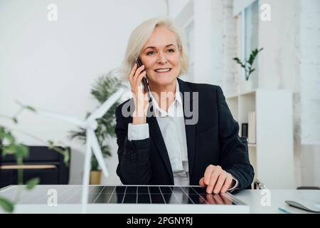 Reife Geschäftsfrau, die mit Solarpaneel sitzt und am Schreibtisch mit einem Mobiltelefon spricht Stockfoto