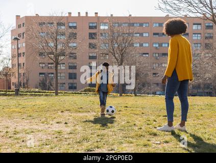 Ein Junge, der mit seiner Mutter Fußball spielt, im Park an einem sonnigen Tag Stockfoto