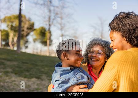 Glückliche Seniorin, die ihren Enkel trägt und mit ihrer Mutter im Park Spaß hat Stockfoto