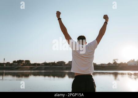 Junger Mann, der mit erhobenen Armen am See steht Stockfoto