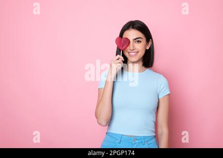 Glückliche Frau, die einen herzförmigen Lutscher über das Auge hält, vor pinkfarbenem Hintergrund Stockfoto