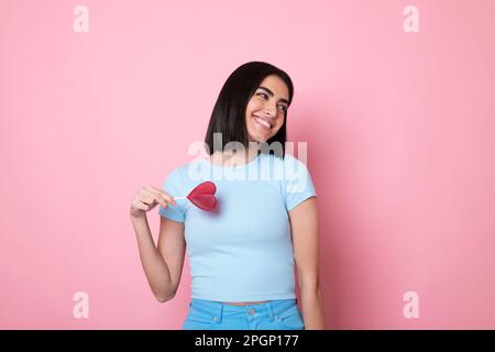Glückliche junge Frau, die einen herzförmigen Lutscher über das Auge hält, vor pinkfarbenem Hintergrund Stockfoto