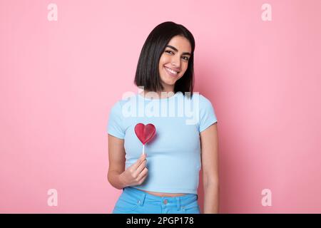 Lächelnde junge Frau mit herzförmigem Lutscher vor pinkfarbenem Hintergrund Stockfoto